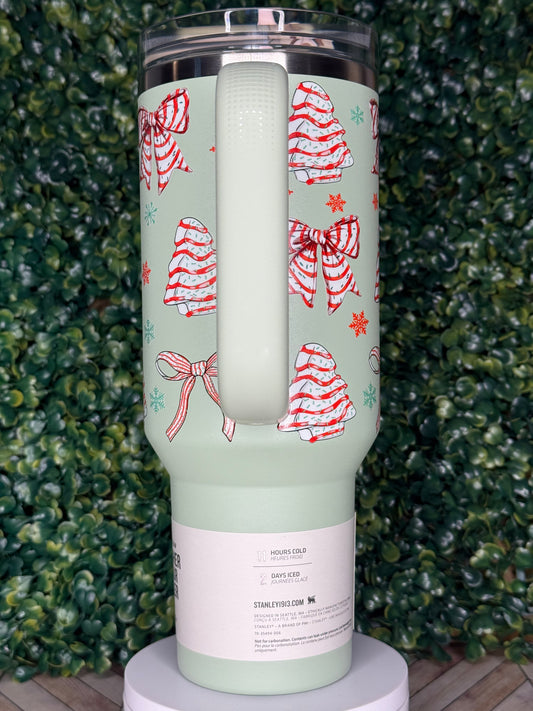 A green insulated tumbler with a festive pattern of red and white bows and snowflakes on it, placed on a surface with green foliage.