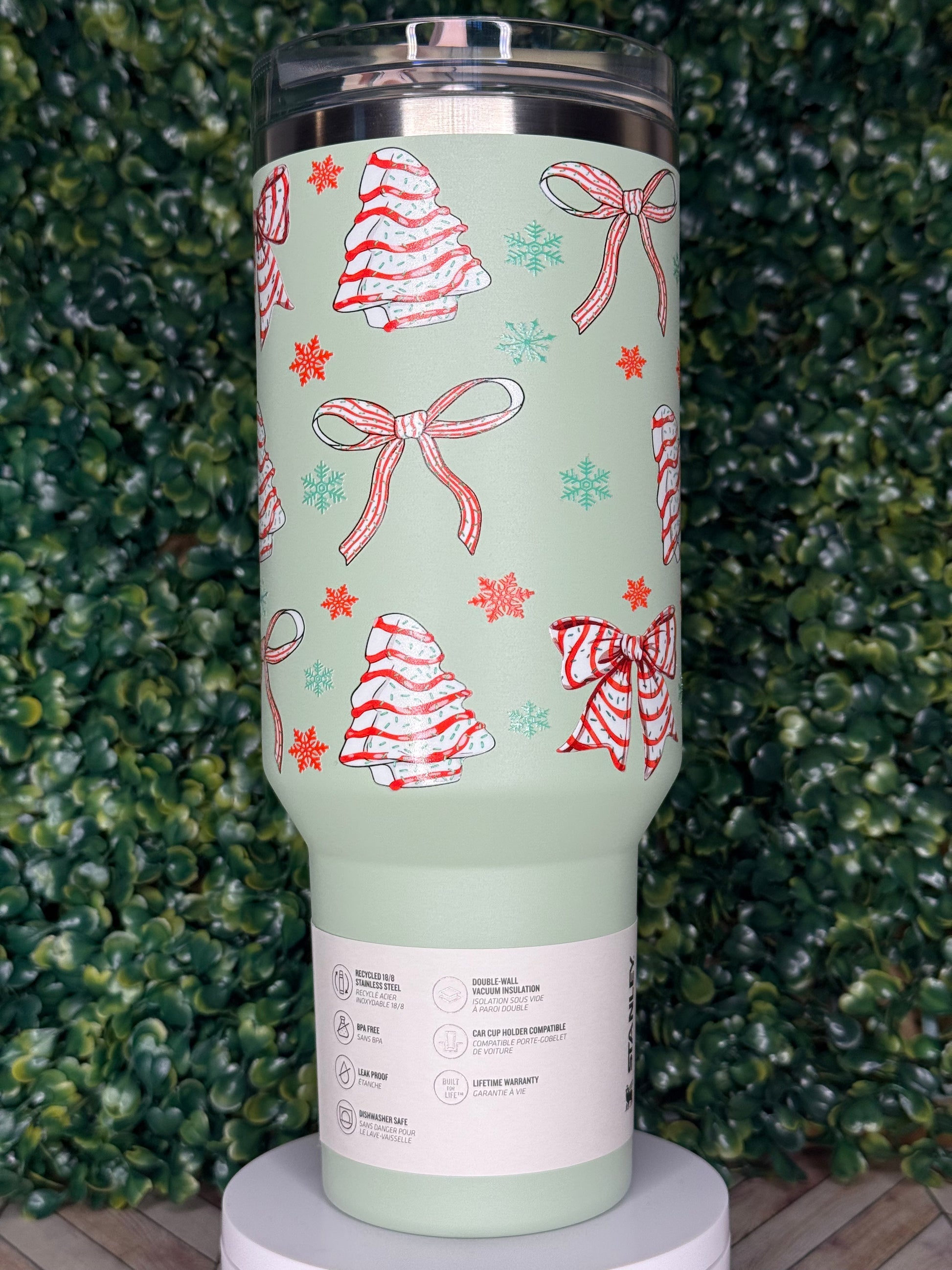 A green tumbler with a festive design of Christmas trees and snowflakes printed on it, placed on a surface with green foliage.