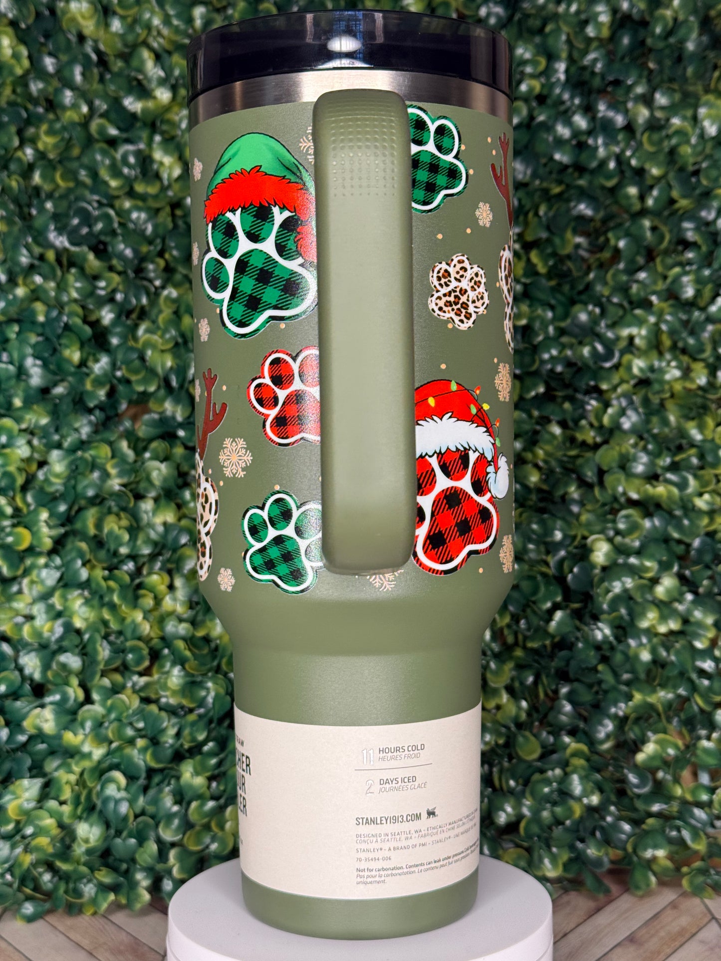A green thermos or thermos-like container with a pattern of paw prints and snowflakes on it, placed on a bed of green leaves.