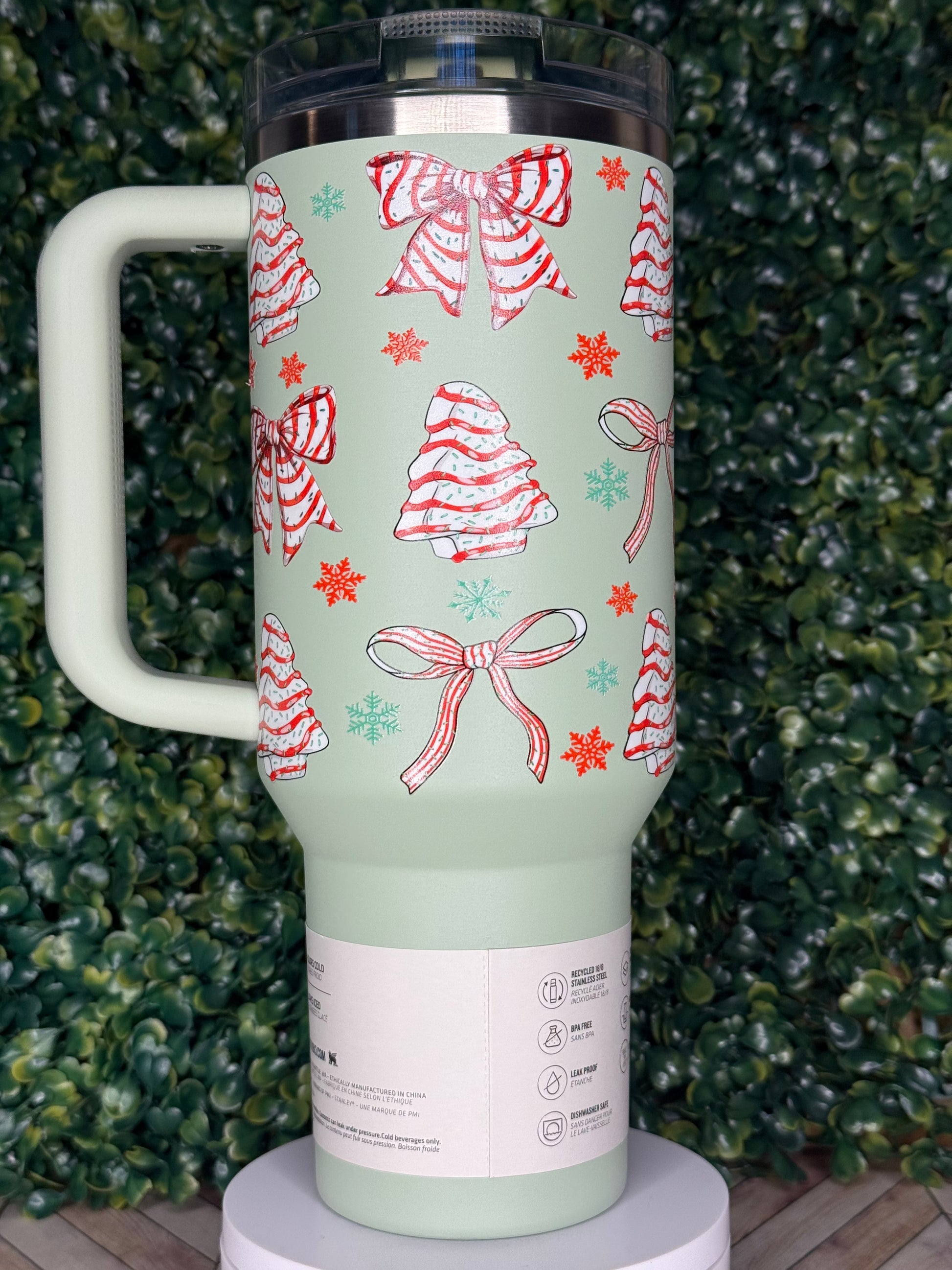 A green tumbler with a festive design of red and white bows and snowflakes on it, placed on a surface with green foliage.