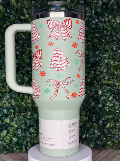 A green tumbler with a festive design of red and white bows and snowflakes on it, placed on a surface with green foliage.
