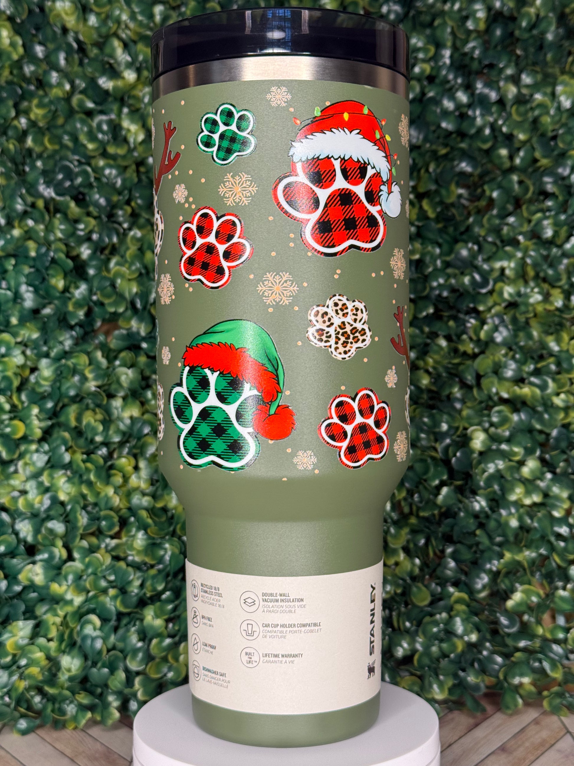A green insulated tumbler with a pattern of paw prints and snowflakes on it, placed on a bed of green leaves.